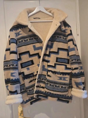 Beige teddyjacka med blått mönster - Snygg teddyjacka i beige med blått och svart geometriskt mönster. Jackan har stor krage, två stora fickor framtill och mysigt teddyfoder. Perfekt för kalla dagar när du vill sticka ut lite extra.