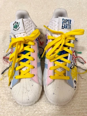 Adidas Super Earth sneakers med brodyr - Unika Adidas Super Earth sneakers i vitt med färgglada broderade blommor och fransar på sidorna. Skorna har gula skosnören, rosa och blå detaljer samt Super Earth-logga på plösen och hälen. Klassisk snäcktå och tillverkade i läder och textil.