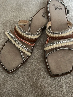 Beige sandaler med pärldetaljer Alma Blue - Snygga beige sandaler från Alma Blue med fyrkantig tå och flera remmar dekorerade med pärlor, flätade band och små bollar i vitt och brunt. Sulan är i syntetmaterial och remmarna har coola detaljer i silver och orange. Perfekta för en trendig sommarlook!