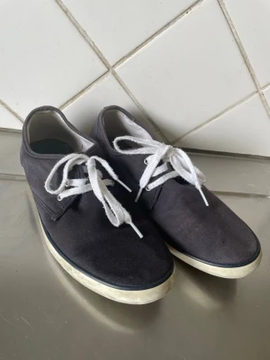Mörkblå sneakers med vita sulor - Snygga mörkblå sneakers med klassisk låg profil och vita snören. Skorna har en vit gummisula med diskret mönster undertill och ovandel i textil. Perfekta för en clean och enkel stil.