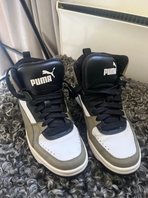Puma sneakers i vitt och olivgrönt - Snygga Puma sneakers i vitt, olivgrönt och svart med högt skaft. Skorna har klassisk snörning, svart plös med Puma-logga och detaljer i syntetläder. Perfekta för dig som gillar streetwear och vill ha en sportig look.