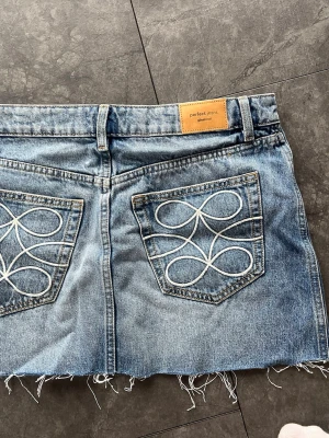 Kort blå jeans skjol med broderade bakfickor - Kort blå jeanskjol från gina med rå, fransig nederkant och snygga broderade mönster på bakfickorna. Kjolen har klassiska fickor fram och bak, bälteshällor och knappstängning. Perfekt för dig som gillar en avslappnad och trendig look. midjemått 38cm och längd 32cm 