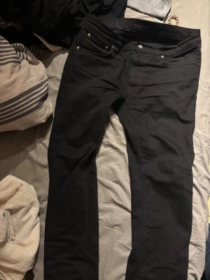 Svarta jeans från Nudie Jeans - Säljer ett par svarta jeans från Nudie Jeans med klassisk femficksmodell och raka ben. Jeansen har en stilren look och är tillverkade i ett mjukt denimtyg som sitter snyggt. Perfekta för dig som gillar minimalistisk och tidlös stil.