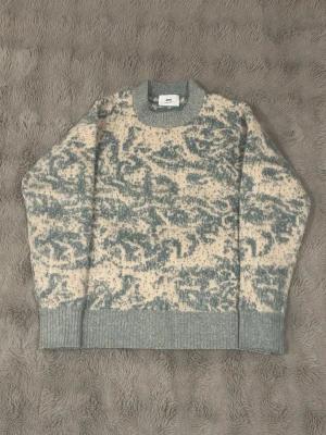 Stickad tröja från Ami i blått och beige - Säljer en stickad tröja från Ami med unikt mönster i blått och beige. Tröjan har rund hals, ribbade muddar och är långärmad. Materialet är mjuk ull och designen är loose fit, perfekt för dig som gillar avslappnad stil.