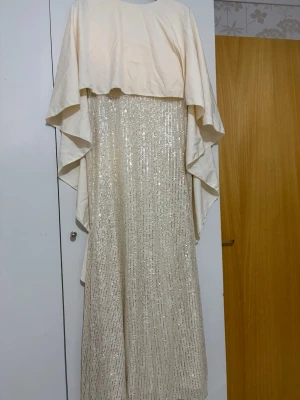 Lång glittrig beige klänning - Elegant långklänning i  beige med glittriga paljettdetaljer på kjolen och ett flytande lager över axlarna. Klänningen har en lös passform och är perfekt för speciella tillfällen. Materialet känns mjukt och skimrande, vilket ger en lyxig känsla.
