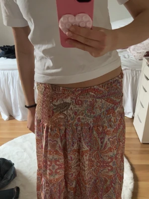 Mönstrad långkjol från Lululia - Säljer en bohemisk långkjol från Lululia/Marta med färgglatt paisleymönster i rosa, orange och beige toner. Kjolen har smock i midjan för skön passform och är gjord i ett lätt och luftigt material, perfekt för varma dagar. Kjolen är använd 2 gånger, Nypris:599 kr💞