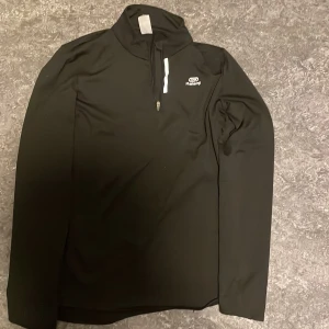 Svart långärmad träningströja Kalenji - Svart långärmad träningströja från Kalenji med halv dragkedja framtill och reflekterande detalj vid dragkedjan. Tillverkad i ett stretchigt funktionsmaterial som passar perfekt för löpning eller annan träning.