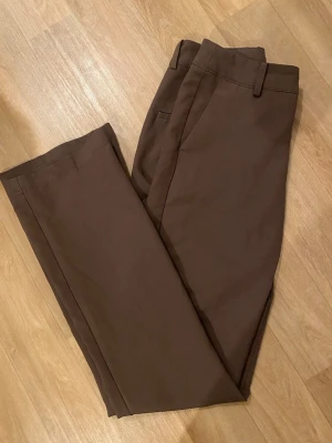 Bruna kostymbyxor från BikBok - Snygga bruna kostymbyxor från BikBok i rak modell. Byxorna har hällor för skärp, sidofickor och är tillverkade i ett mjukt tyg som känns bekvämt. Perfekta för en stilren look och lätta att matcha med olika toppar.