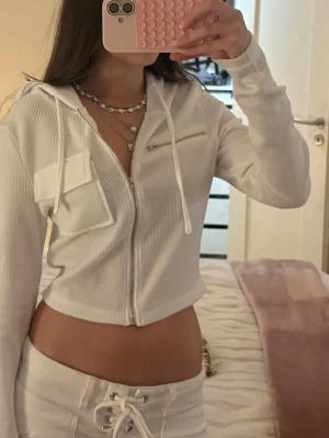 Vit cropped hoodie med dragkedja - Säljer en vit cropped hoodie med dragkedja framtill, ribbad struktur och huva med snören. Hoodien har en bröstficka med lock och en liten dragkedjeficka. Perfekt för en chill och trendig look.