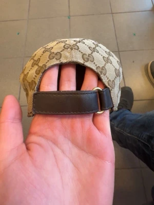 Gucci keps - Säljer en beige keps från Gucci med det klassiska GG-monogrammönstret och justerbar rem i brunt skinn baktill. Kepsen har böjd skärm och är perfekt för dig som vill ha en stilren accessoar med lyxig känsla.