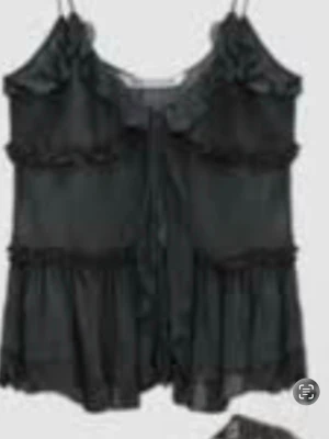 Svart spetsbody med volanger - Säljer en svart body med tunna axelband, volangdetaljer och transparent tyg. Bodyn har en söt rosett framtill och är dekorerad med spets för en extra feminin touch. Perfekt för dig som vill ha något både snyggt och bekvämt.