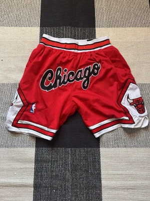 Chicago Bulls basketshorts röd - Säljer ett par röda Chicago Bulls basketshorts från Just Don med svart och vit text, samt Bulls-logga på sidorna. Shortsen har elastisk midja, vita och svarta detaljer och är tillverkade i mesh-liknande polyester. Perfekta för dig som gillar streetwear och basket.