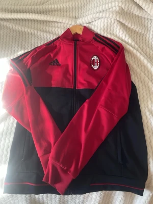AC Milan träningsjacka från Adidas - Röd och svart träningströja från Adidas med AC Milan-logga på bröstet. Tröjan  har hel dragkedja, ståkrage och svarta detaljer på axlarna. Perfekt för fotbollsfans som vill ha en sportig look. Tillverkad i ett mjukt och funktionellt material.