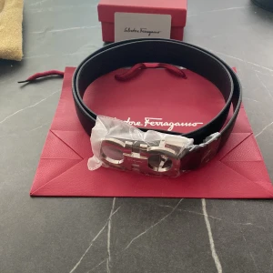 Svart bälte från Salvatore Ferragamo - Stilrent svart bälte från Salvatore Ferragamo med ikoniskt silverfärgat spänne. Bältet har en klassisk design och är tillverkat i slätt skinn. Perfekt accessoar för att lyfta vilken outfit som helst.