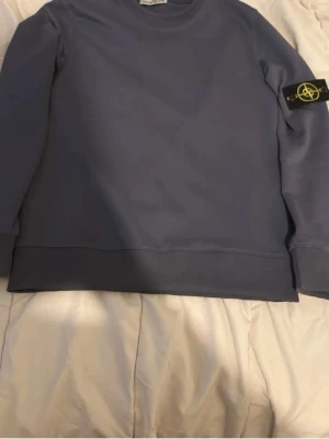 Blå stickad tröja från Stone Island - Säljer en stilren blå stickad tröja från Stone Island med klassisk rund hals och den ikoniska logotypen på vänster ärm. Tröjan har ribbade muddar och nederkant för en snygg passform. Perfekt för dig som gillar streetwear och enkel design.