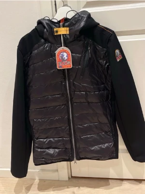 Svart pufferjacka från Parajumpers - Snygg svart pufferjacka från Parajumpers med huva och dragkedja framtill. Jackan har quiltad front, släta ärmar och en bröstficka med dragkedja. Märkets patch sitter på ärmen och detaljer i gult vid halsen ger extra stilpoäng.
