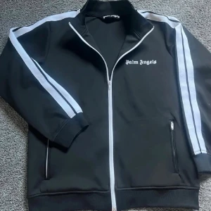 Ny Svart ziptröja Palm Angels - Säljer en svart ziptröja från Palm Angels med vita ränder längs ärmarna och logga på bröstet. Tröjan har hög krage, dragkedja framtill och två fickor med dragkedja. Perfekt för en sportig och trendig look.