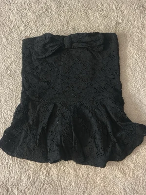 Svart spets tubtopp med rosett - Säljer en svart axelbandslös tubtopp i spets med en stor rosett framtill och volang nedtill. Toppen har en feminin och elegant look med fina spetsdetaljer och är perfekt för dig som vill sticka ut lite extra.
