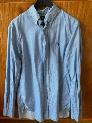 Blå långärmad skjorta från Polo Ralph Lauren - Klassisk blå skjorta från Polo Ralph Lauren med diskret rutmönster och broderad logga på bröstet. Skjortan har button-down krage, långa ärmar och knappar framtill. Perfekt till både jeans och chinos för en clean look. Bra skick behöver bara strykas