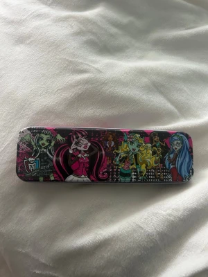 Monster High pennskrin i plåt - Säljer ett färgglatt Monster High pennskrin i plåt med motiv av flera karaktärer på locket. Skrinet är svart, rosa och blått och har plats för pennor och småsaker. Ett blått pennliknande föremål med Monster High-berlock ingår.