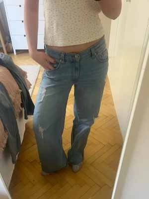 Ljusblåa vida jeans med slitningar - Säljer ett par ljusblåa jeans med breda ben och slitna detaljer på framsidan. Jeansen har hög midja, klassiska fem fickor och råa kanter vid bensluten. Perfekta för en avslappnad och trendig look.
