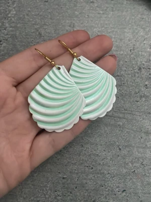 Mintgröna snäckformade örhängen - Unika örhängen i form av snäckskal med mjuka vita och mintgröna nyanser. De har guldfärgade krokar och är tillverkade i ett lätt material som liknar plast eller resin. Perfekta för dig som gillar strandinspirerad stil och vill sticka ut med coola accessoarer.