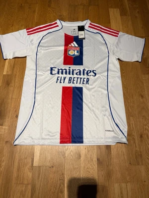 Lyon 25/26 home kit endrick #9 - Snygg vit Olympique Lyon matchtröja från Adidas med röda och blå detaljer, klubbmärke på bröstet och Emirates Fly Better-tryck. Röd och blå rand framtill, Endrick och nummer 9 på ryggen. Tillverkad i lätt och ventilerande Aeroready-material.