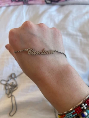 Silverfärgat namnhalsband Carolina - Snyggt halsband i silverfärg med namnet 'Carolina” i skrivstil. Kedjan är tunn och smidig, och namndetaljen är i metall. Perfekt accessoar för dig som vill ha ett personligt smycke.