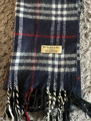 Mörkblå rutig halsduk från Burberry - Klassisk halsduk från Burberry i mörkblått med vitt och rött rutmönster. Tillverkad i 100% lammull och har fransar i ändarna. Perfekt accessoar för att lyfta din outfit med en tidlös och lyxig känsla.