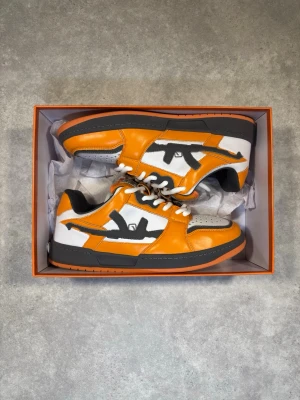 Lost Boys orange låga sneakers - Snygga sneakers från Lost Boys i orange, vitt och svart. Skorna har en låg silhuett, vita snören och svart sula. Sidopanelerna har en unik svart grafisk detalj och ovandelen är i en mix av läder och textil. Perfekta för dig som vill sticka ut.