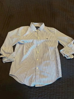 Randig skjorta från Tommy Hilfiger - Klassisk vit och blå randig skjorta från Tommy Hilfiger med lång ärm och knappar framtill. Skjortan har en liten broderad logga på bröstet och är tillverkad i bomull. Perfekt för en stilren och avslappnad look.