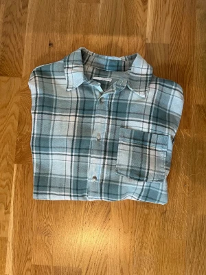 Grönrutig skjorta i bomull - Säljer en grön och vit rutig skjorta med klassisk krage och bröstficka. Skjortan har långa ärmar och knäppning framtill. Perfekt för dig som gillar en avslappnad och stilren look.