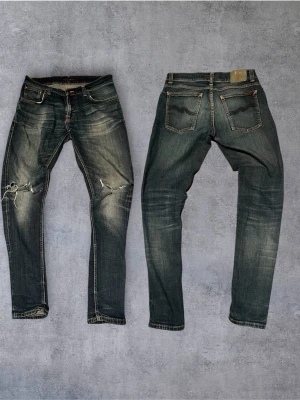 Vintage Nudie Jeans - Säljer ett par blå jeans från Nudie Jeans med slitningar och hål på knäna. Jeansen har klassisk femficksmodell, orangea sömmar och en snygg tvättad look. Perfekta för dig som gillar en avslappnad och trendig stil.