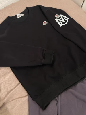 M*ncler sweatshirt - Säljer en svart sweatshirt från M*ncler med klassisk rund halsringning. Tröjan har Mo*cler-logga på bröstet och en stor vit Moncle*-dekal på ärmen. Tillverkad i mjukt material och har ribbade muddar vid ärmslut och nederkant. Perfekt för en clean och stilren look.