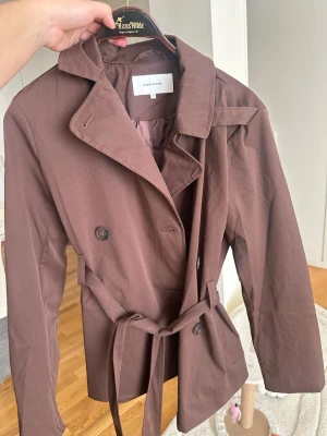Brun trenchcoat från BubbleRoom - Snygg brun trenchcoat från BubbleRoom med klassisk dubbelknäppning, bred krage och knytskärp i midjan. Jackan har långa ärmar och är tillverkad i ett slätt tyg som ger en stilren look. Perfekt för dig som vill ha en trendig och tidlös jacka. Oanvänd helt nytt