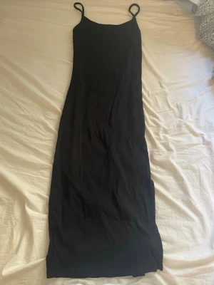 Svart långklänning med tunna band - En enkel och stilren svart långklänning med tunna axelband och rak passform. Klänningen har en minimalistisk design och är perfekt för dig som gillar en clean look. Materialet känns mjukt och följsamt, vilket gör den bekväm att bära.