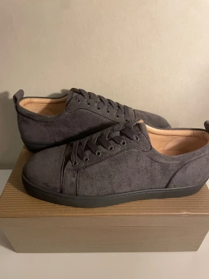 Gråa sneakers från Christian Louboutin - Snygga gråa sneakers i mocka från Christian Louboutin med klassisk röd sula och diskret logga undertill. Skorna har lågt skaft, matchande gråa skosnören och en stilren design som passar till det mesta. Insidan är ljusbeige och sulan är i gummi.