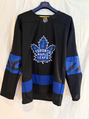 Toronto Maple Leafs Nylander #88 - Snygg svart hockeytröja från Fanatics med Toronto Maple Leafs-logga i blått och vitt på bröstet. Stora blå ränder på ärmar och midja, samt namn och nummer 88 'Nylander' på ryggen. Tillverkad i lätt och ventilerande syntetmaterial, perfekt för hockeyfans.