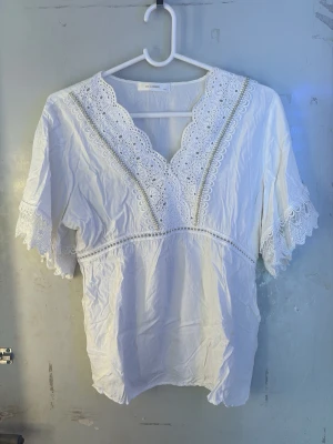 Vit spetsblus med v-ringning - Söt vit blus med v-ringning och dekorativa spetsdetaljer längs halsringningen och ärmsluten. Blusen har korta ärmar med spetskant och lätt rynkad midja för en feminin siluett. Tillverkad i ett tunt, luftigt material.