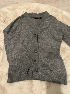 Grå stickad kofta med knappar - Mysig grå stickad kofta med tre stora knappar framtill och två fickor. Koftan har ribbade muddar vid ärmslut och nederkant samt en klassisk V-ringning. Perfekt att slänga på sig när det är kyligt ute.