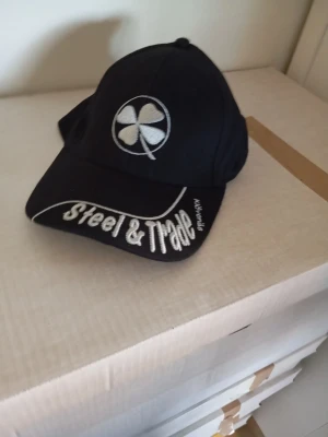 Svart keps Steel & Trade - Säljer en svart keps från Steel & Trade med vit broderad fyrklöver på framsidan och texten 'Steel & Trade' broderad på skärmen. Kepsen har klassisk böjd skärm och justerbar baksida. Perfekt för dig som gillar streetwear och vill sticka ut.