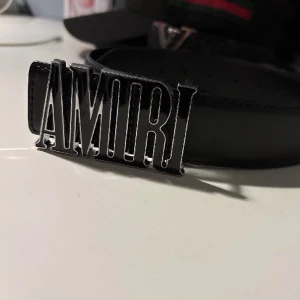Svart bälte från Amiri med metallspänne - Snyggt svart bälte från Amiri med ett stort metallspänne där märkesnamnet står i silver. Bältet har en clean look och passar perfekt till jeans eller byxor för en trendig stil.