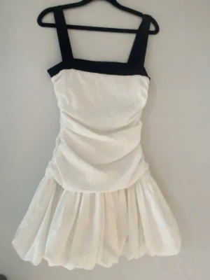 Vit kortklänning med svarta axelband - Säljer en vit kortklänning med breda svarta axelband och fyrkantig ringning. Klänningen har en puffig kjol och rynkade detaljer i midjan. Perfekt för dig som vill sticka ut med en unik och trendig look.