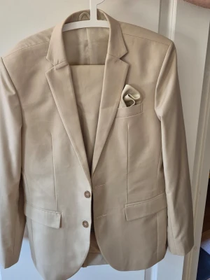 Beige kostym med kavaj och byxor - Stilren beige kostym med matchande kavaj och byxor. Kavajen har två knappar, klassisk krage och en bröstficka med näsduk. Kostymen har en elegant och tidlös design, perfekt för dig som vill ha en clean och modern look.