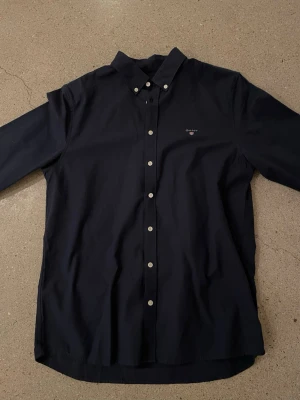 Mörkblå skjorta från Gant - Snygg mörkblå skjorta från Gant med klassisk button-down krage och vita knappar. Skjortan har lång ärm och diskret Gant-logga broderad på bröstet. Perfekt för dig som gillar stilrena och tidlösa plagg.