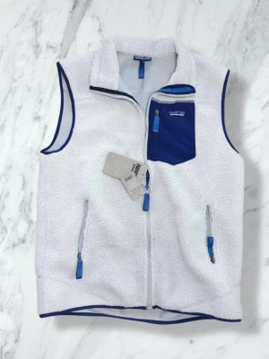 Patagonia väst, oanvänd - Snygg beige fleeceväst från Patagonia med blå detaljer och dragkedjor. Perfekt för lager-på-lager och chill stil. Västen är helt oanvänd och lapparna sitter kvar. Hör gärna av dig vid frågor!