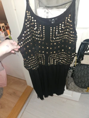 Svart klänning med guldpaljetter H&M - Svart kortklänning från H&M med guldiga paljettdetaljer i geometriskt mönster på överdelen. Klänningen är ärmlös och har en lös passform upptill. Perfekt för dig som vill sticka ut med lite extra bling. Köpt på H&M för över 20 år sen men är i fantastiskt skick 