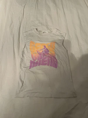 Vit ärmlös t-shirt från H&M - Säljer en vit ärmlös t-shirt från H&M med färgglatt tryck i orange och lila på bröstet där det står 'Fresh Vibes Ahead'. T-shirten har rund halsringning och är gjord i mjuk bomull, perfekt för en avslappnad och cool stil.