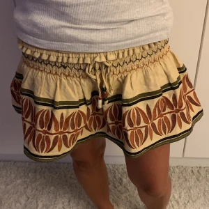 Beige kortkjol med snörning och mönster - Supersöt beige kortkjol med elastisk midja och snörning med pärlor. Kjolen har ett brunt och grönt grafiskt mönster längs kanten och volang för extra flow. Perfekt för dig som gillar boho-vibe och vill sticka ut med coola detaljer.