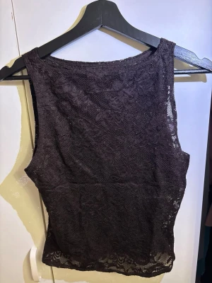 Mörkbrun spetslinne med blommönster - Säljer en mörkbrun topp i genombruten spets med blommönster. Toppen har en rund halsringning och är figurnära i modellen. 